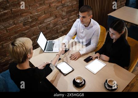 Drei Profis führen an einem Holztisch in einer gemütlichen Coffee-Bar mit moderner Technologie ein ernsthaftes Geschäftsgespräch. Stockfoto