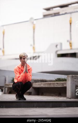 Dieses Bild zeigt eine junge Frau in einem leuchtend orangefarbenen Jackett, die während ihres morgendlichen Joggs eine friedliche Pause mit einer Meditationspose einlegt. Stockfoto