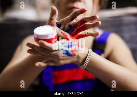 Eine Künstlerin setzt sich mit ihrer Kreativität auseinander und mischt leuchtende rote und blaue Farben mit ihren Händen, umgeben von ihren Kunstwerkzeugen. Stockfoto