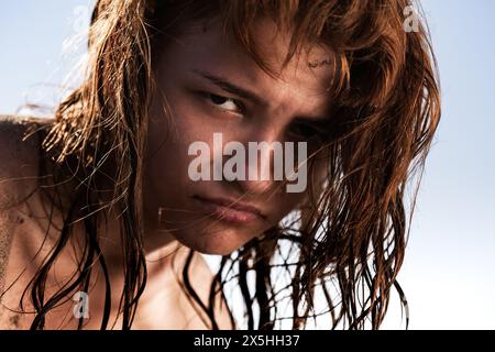 Dieses eindrucksvolle Bild fängt eine junge Frau mit nassen, verworrenen Haaren ein, deren Blick intensiv auf einen klaren blauen Himmel gerichtet ist. Das natürliche Licht betont die Textur Stockfoto