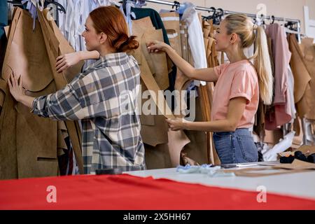 Zwei erfolgreiche Modedesignerinnen, die in einer Textilfabrik arbeiten, Stoff wählen und reden. Zwei Damen, die ein paar Kleidungsstücke besorgen Stockfoto