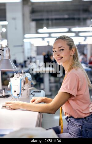 Junger netter kaukaischer blonder Textilfabrikarbeiter auf der Produktionslinie mit moderner Nähmaschine in der Werkstatt. Fleißige Frau, die mit dem Nähen beschäftigt ist oder Stockfoto
