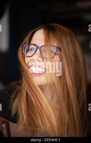 Eine fröhliche junge Frau mit Brille und langen blonden Haaren lächelt breit, ihre Haare wehen beiläufig, während sie einen fröhlichen Moment genießt. Stockfoto