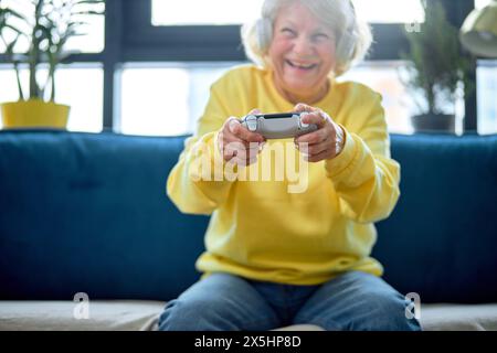 Lustige europäische Seniorin mit grauen Haaren und Joystick spielen Online-Spiel in der Freizeit im Innenraum, allein. Ältere Menschen und moderne Geräte, Rentner und moderne Technologien Konzept Stockfoto