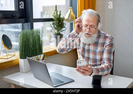 Senior 70er-Mann sitzen entspannen Sie sich in einem gemütlichen Zimmer, surfen Sie im Internet auf einem modernen Laptop und hören Sie Musik mit Smartphone und Headset. Reifer Großvater Ruhen Sie sich zu Hause aus Video surfen Sie drahtlos im Internet auf einem Laptop Stockfoto