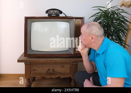 Älterer Mann in blauem Polohemd sitzt auf dem Boden vor dem alten analogen Retro-Fernseher, Fernseher, weißem Bildschirm Mockup für Designer, Fernsehen schauen, ältere Menschen Stockfoto