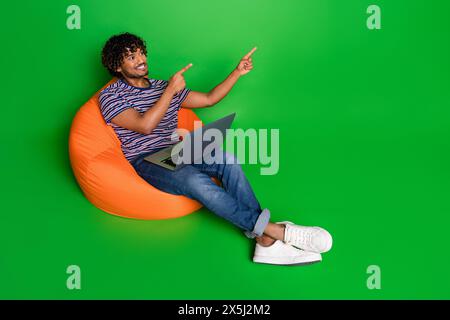 Foto eines jungen Studentinnen, der Laptop für die Arbeit benutzt, wenn er auf Pouf Point Fingers liegt Website-Design isoliert über grünem Hintergrund Stockfoto