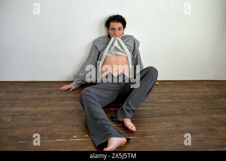 Junger, gutaussehender Mann, der in seinen Pullover beißt. Blick auf die Kamera. Auf dem Boden liegend, in voller Länge. Guy hat zu Hause Spaß Stockfoto
