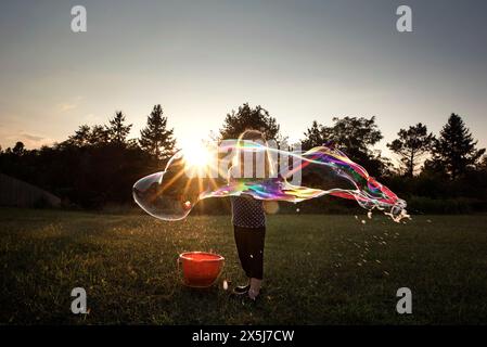 Ein kleines Mädchen, das bei Sonnenuntergang auf einem grasbewachsenen Feld eine riesige bunte Blase bläst Stockfoto