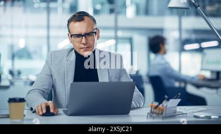 Modernes Büro: Porträt eines erfolgreichen Geschäftsmannes mittleren Alters mit Anzug und Brille, der an einem Laptop an seinem Schreibtisch arbeitet. Seriöse, Selbstbewusste Und Stilvolle Unternehmensleitung Mit Computer. Vorderansicht Aufnahme Stockfoto