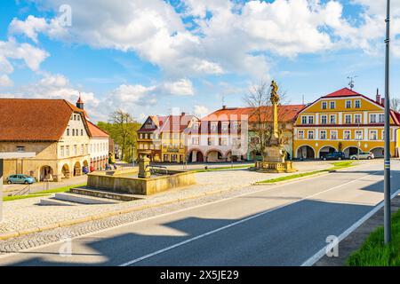 Ein malerischer Blick auf einen friedlichen Platz in Zacler, Tschechische Republik. Farbenfrohe Gebäude mit klassischer europäischer Architektur säumen die Straße. Ein wunderschöner Brunnen mit einer komplizierten Statue befindet sich in der Mitte. Der klare blaue Himmel lässt einen sonnigen Tag vermuten. Eine gepflegte asphaltierte Straße führt durch den Platz. Stockfoto