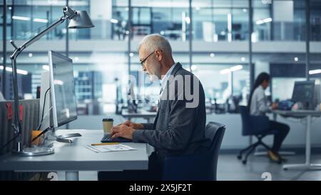 Modernes Büro: Porträt eines erfolgreichen bärtigen Geschäftsmannes mittleren Alters, der an seinem Schreibtisch an einem Laptop arbeitet. Lächelnder Unternehmensmitarbeiter. Multiethnische Arbeitsumgebung mit zufriedenen Profis. Stockfoto