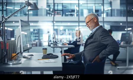 Senior White männlicher Mitarbeiter im Unternehmensbüro arbeitet am Computer und fühlt plötzliche Rückenschmerzen. Der Buchhalter fühlt sich krank wegen der ganzen Arbeit, Stress, Rückenschmerzen Stockfoto