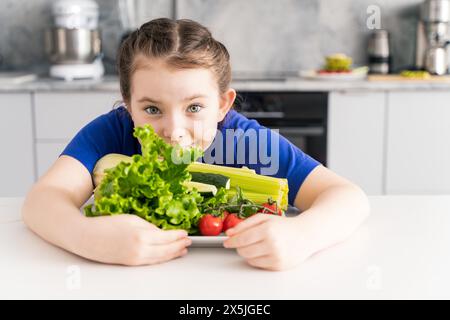 Schönes und glückliches kleines Mädchen in einem blauen T-Shirt mit einem Teller mit frischem Gemüse auf dem Hintergrund einer grauen Küche. Hochwertige Fotos Stockfoto