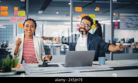 Zwei Happy Young Professionals im Büro: Schwarze Geschäftsfrau und hispanische Geschäftsmann tanzen zusammen, lächeln und in die Kamera schauen, am Schreibtisch sitzen. Spezialisten arbeiten in Creative Office, feiern Erfolge Stockfoto
