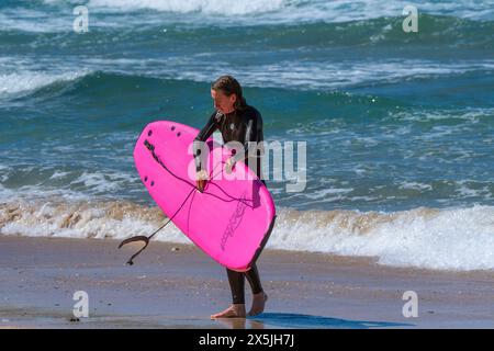 Eine Surferin, die ihr Surfbrett trägt und am Fistral Beach in Newquay in Cornwall in Großbritannien aus dem Meer wandert. Stockfoto