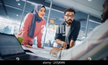 Diversity Office Big Table Meeting: Muslimische Geschäftsfrau trägt Hijab und hispanische männliche Unternehmerin arbeiten mit Dokumenten, Talk, Brainstorming. Fachleute im E-Commerce-Projekt. Niederländischer Winkel Stockfoto