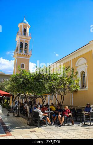 Argostoli, Griechenland, Kefalonia, Menschen in einem von Bäumen beschatteten Café im Freien und einer blauen Kuppel gelben griechischen Kirche. (Nur Für Redaktionelle Zwecke) Stockfoto