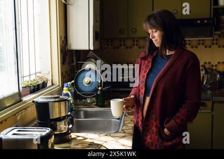 Eine erwachsene Latina mit rotem Pullover steht mit geschlossenen Augen vor dem Küchenfenster, ruhig und nachdenklich, während sie Kaffee trinkt. Stockfoto