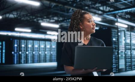 Weibliche Reife multirassische E-Business-Unternehmerin Schau weg und hält Laptop im Data Center Server Abendraum. Technologie Web Services Cloud Computing und SAAS-Konzept Stockfoto