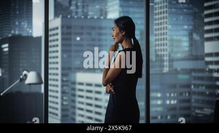 Erfolgreiche Geschäftsfrau in stilvollem Kleid, die an einem Laptop arbeitet und aus dem Fenster in Big City blickt. Selbstbewusste CEO analysiert Finanzprojekte. Marketingkampagne für Manager bei der Arbeitsplanung. Stockfoto