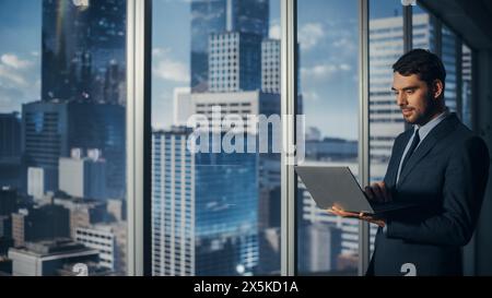 Geschäftsmann steht im modernen Büro, mit Laptop-Computer, neben dem Fenster mit Blick auf Big City mit Wolkenkratzer. Erfolgreicher CEO oder Finanzmanager, der Investitionsprojekte plant. Stockfoto