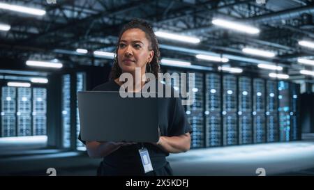 IT-Techniker für multirassische Rechenzentren, der mit einem Laptop im Server-Rack-Korridor steht. Sie überprüft etwas visuell, während sie wegschaut. Nachtbürokonzept Stockfoto