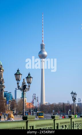 Der Berliner Fernsehturm steht hoch vor einem klaren blauen Himmel, flankiert von Vintage-Straßenlaternen, und Besucher genießen die Aussicht. Stockfoto