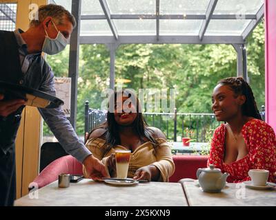 Kellner in Gesichtsmaske serviert Latte an lächelnden Frauen im Café Stockfoto