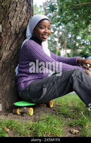 Großbritannien, London, Junge Frau im Hijab, die auf Skateboard am Baumstamm sitzt Stockfoto