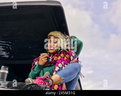 Frau, die auf dem Dach des Autos sitzt und aus einem isolierten Getränkekontainer trinkt Stockfoto