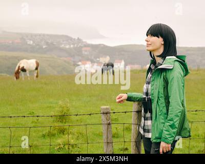 Großbritannien, Frau, die im Grasfeld steht, bei Pferden, die im Hintergrund grasen Stockfoto