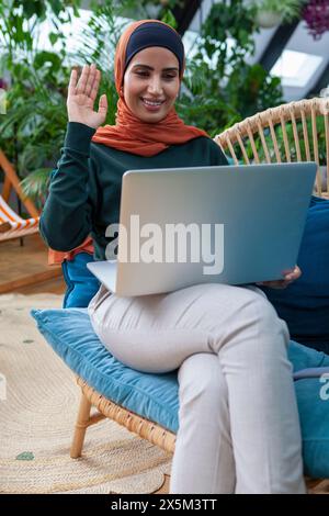 Lächelnde Frau mit Kopftuch und Videoanruf über Laptop zu Hause Stockfoto