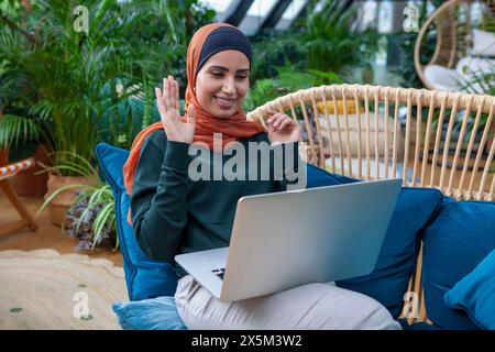 Lächelnde Frau mit Kopftuch und Videoanruf über Laptop zu Hause Stockfoto