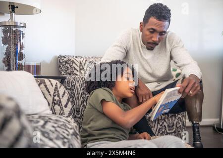 Mann mit Beinprothese liest Buch mit Sohn auf Sofa Stockfoto