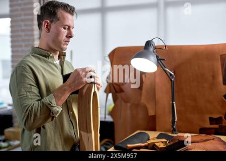 Ehrgeiziger junger Handwerker, der mit Lederstitklemme arbeitet. Nahaufnahme Seitenansicht. Ledernähausrüstung, Mann, der mit Nadel und Faden arbeitet, Brieftasche für Kunden herstellt Stockfoto
