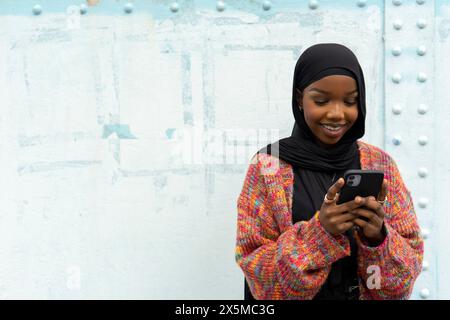 Lächelnde junge Frau im Hijab, die auf das Smartphone blickt Stockfoto