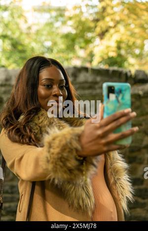 Lächelnde schwangere Frau im Mantel, die im Herbst Selfie im Park macht Stockfoto