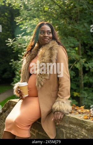 Lächelnde schwangere Frau im Mantel, die im Herbst im Park an der Wand sitzt Stockfoto