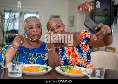 Lächelnde Seniorin mit erwachsener Tochter, die Abendessen isst und Selfie macht Stockfoto