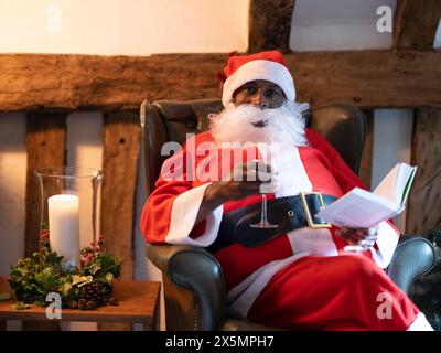 Porträt des Weihnachtsmanns, der Buch liest und Wein im Sessel trinkt Stockfoto