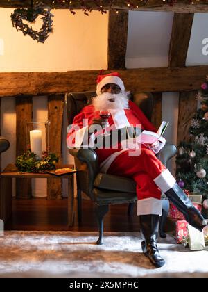 Porträt des Weihnachtsmanns, der Buch liest und Wein im Sessel trinkt Stockfoto