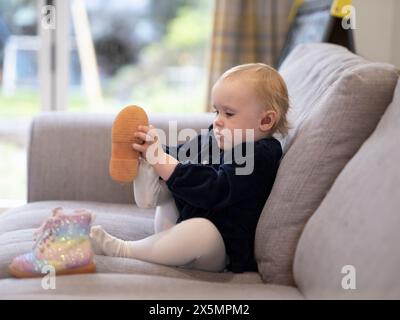 Kleines blondes Mädchen, das auf dem Sofa sitzt und Schuhe anzieht Stockfoto