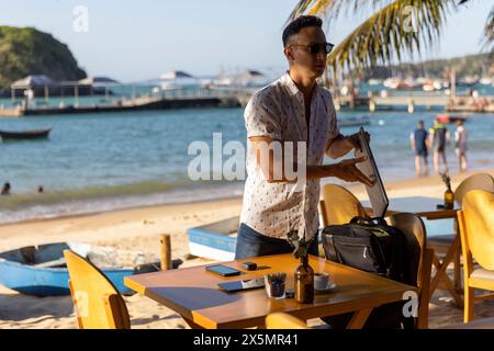 Mann, der mobile Geräte in den Rucksack am Tisch des Strandcafés steckt Stockfoto