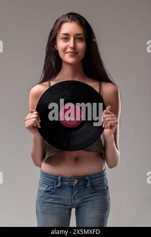 Dünne junge Frau mit flachem Bauch hält eine Schallplatte und steht lässig in Jeans und lächelt sanft Stockfoto