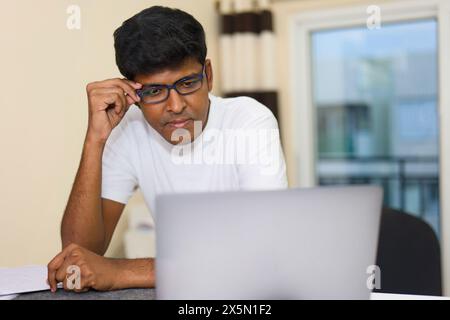 Ein junger Indianer mit Brille arbeitet intensiv an seinem Laptop in einem hell erleuchteten Heimbüro Stockfoto