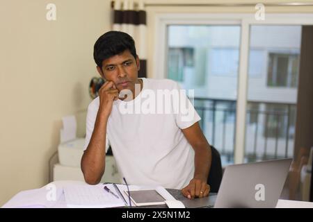 Ein indischer Mann, der nachdenklich und konzentriert aussieht, während er an seinem Laptop an seinem Schreibtisch im Heimbüro arbeitet. Stockfoto