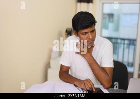 Ein junger Mann sitzt nachdenklich an seinem Schreibtisch in seinem Heimbüro, tief konzentriert, während er an seinem Computer arbeitet. Stockfoto