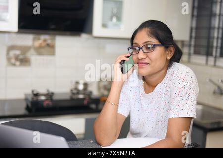 Eine junge Frau lacht, während sie in einer sonnigen Küche mit warmen Tönen am Telefon plaudert. Stockfoto