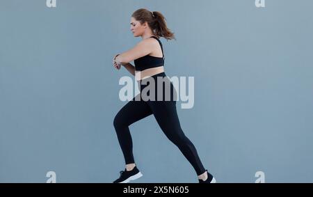 Seitenansicht einer Frau in Übergröße, die mit schwarzer Fitnesskleidung gegen eine graue Wand läuft. Eine junge Frau mit einem großen Körper, der joggt und ihn ansieht Stockfoto